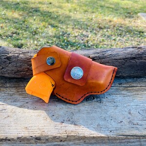 Op de afbeelding: Een handgemaakte leren holster in bruine tinten, met een zilveren muntaccent en een zwarte knoopsluiting. Een oranje pistool is gedeeltelijk zichtbaar. De holster is ontworpen met gestikte randen.