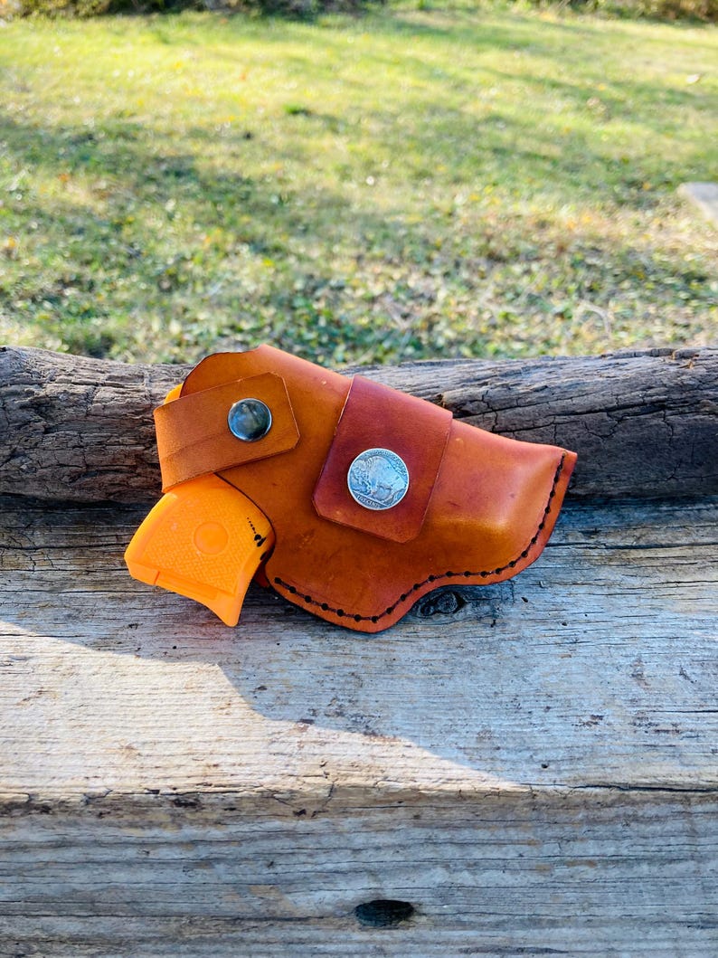 Op de afbeelding: Een handgemaakte leren holster in bruine tinten, met een zilveren muntaccent en een zwarte knoopsluiting. Een oranje pistool is gedeeltelijk zichtbaar. De holster is ontworpen met gestikte randen.