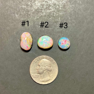 Puede incluir: Tres piedras preciosas de ópalo coloridas, etiquetadas como #1, #2 y #3, se muestran junto a una moneda de veinticinco centavos para comparar el tamaño. Los ópalos exhiben una gama de colores, incluyendo rosa, verde y azul. La moneda está fechada en 1972.