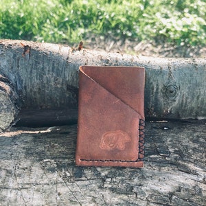 Puede incluir: Una cartera de cuero marr&oacute;n con una silueta de oso grabada en relieve en la parte delantera. La cartera tiene un cierre de solapa y est&aacute; cosida con hilo negro.