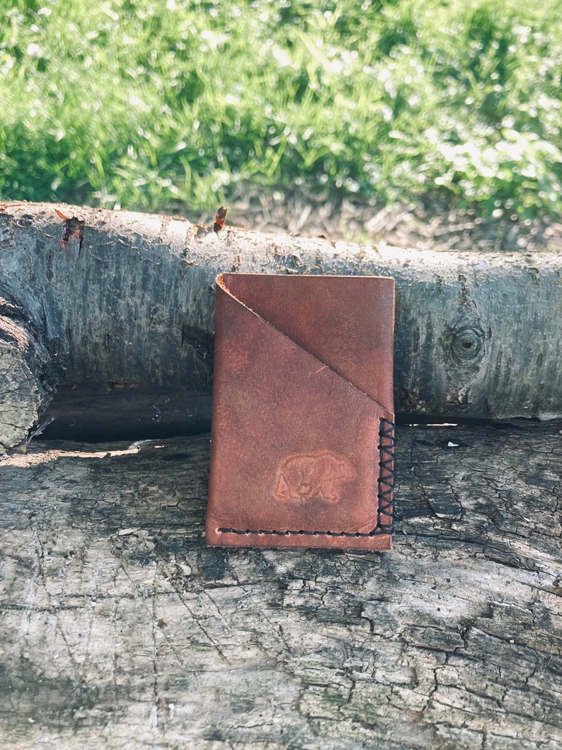 Puede incluir: Una cartera de cuero marr&oacute;n con una silueta de oso grabada en relieve en la parte delantera. La cartera tiene un cierre de solapa y est&aacute; cosida con hilo negro.