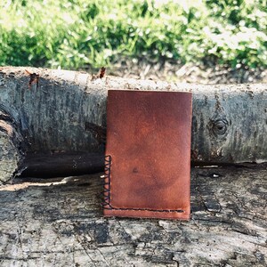 Puede incluir: Una cartera de cuero marr&oacute;n con un borde cosido. La cartera est&aacute; sentada sobre un trozo de madera desgastada.