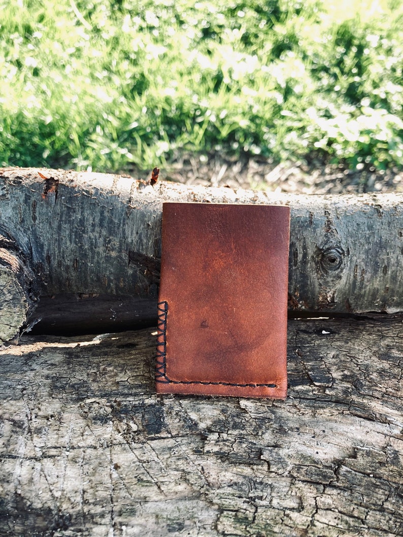 Puede incluir: Una cartera de cuero marr&oacute;n con un borde cosido. La cartera est&aacute; sentada sobre un trozo de madera desgastada.