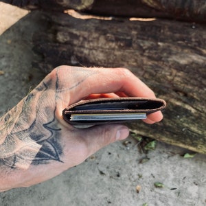 Puede incluir: Una cartera de cuero marr&oacute;n con varias tarjetas en su interior, sostenida en una mano con un tatuaje.