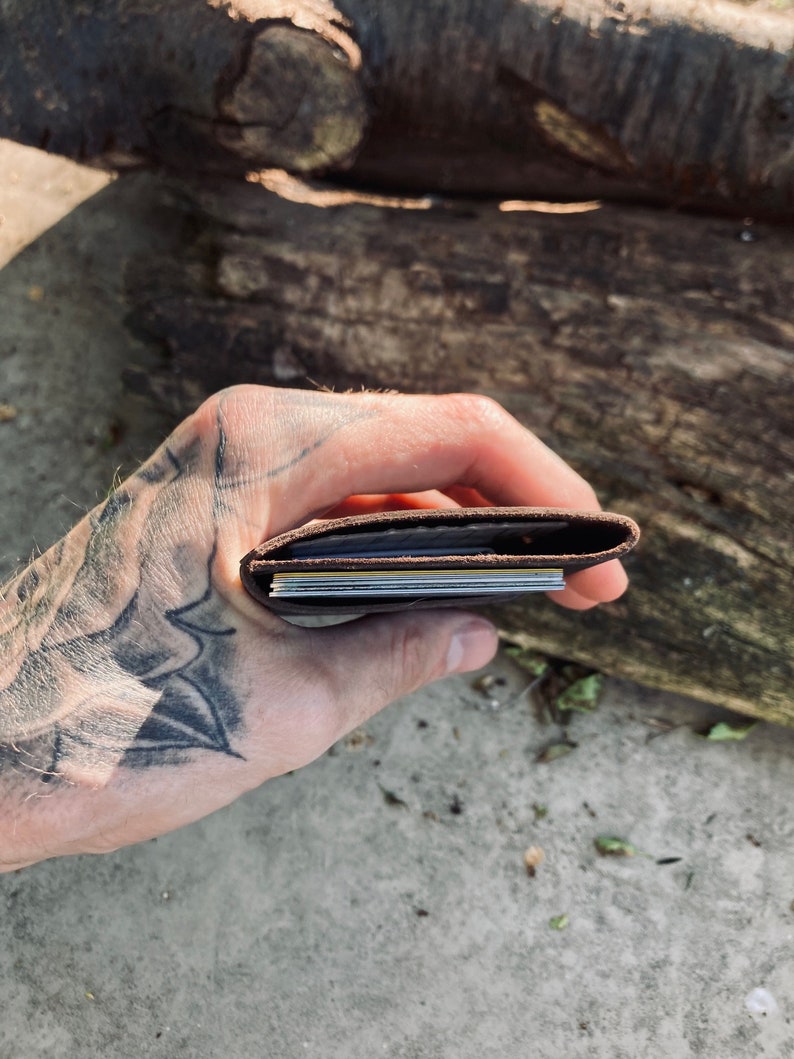 Puede incluir: Una cartera de cuero marr&oacute;n con varias tarjetas en su interior, sostenida en una mano con un tatuaje.