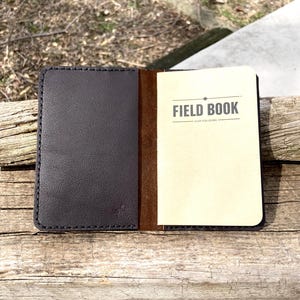 May include: Open leather notebook with the words "FIELD BOOK" printed on the right page. The notebook is open, revealing a cream-colored paper interior. The cover is dark gray with visible stitching.