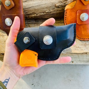 Op de afbeelding: Zwarte leren holster met een zilveren buffelnikkel en druksluiting, met een oranje pistool. Twee extra leren holsters in bruin en bruin zijn zichtbaar op de achtergrond.