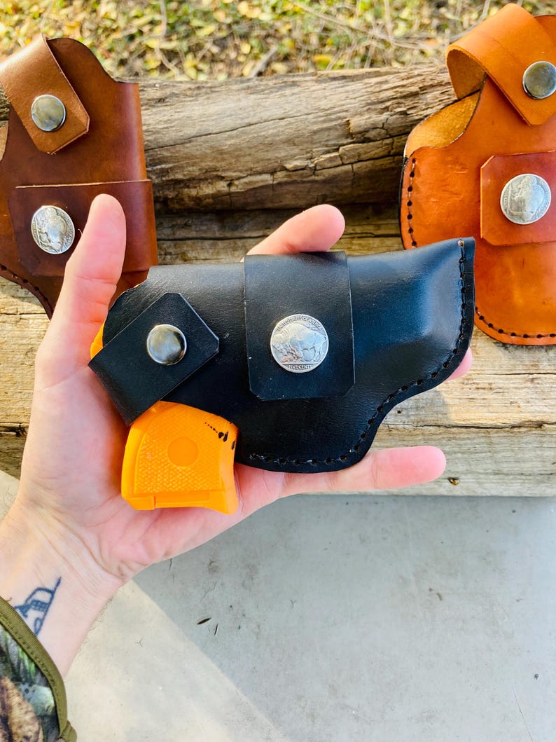 Op de afbeelding: Zwarte leren holster met een zilveren buffelnikkel en druksluiting, met een oranje pistool. Twee extra leren holsters in bruin en bruin zijn zichtbaar op de achtergrond.