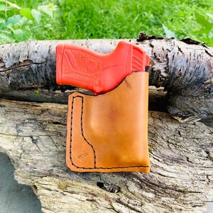 May include: A tan leather holster with black stitching and a red training pistol. The holster has a minimalist design with a stitched logo on the front. The pistol is visible inside the holster.