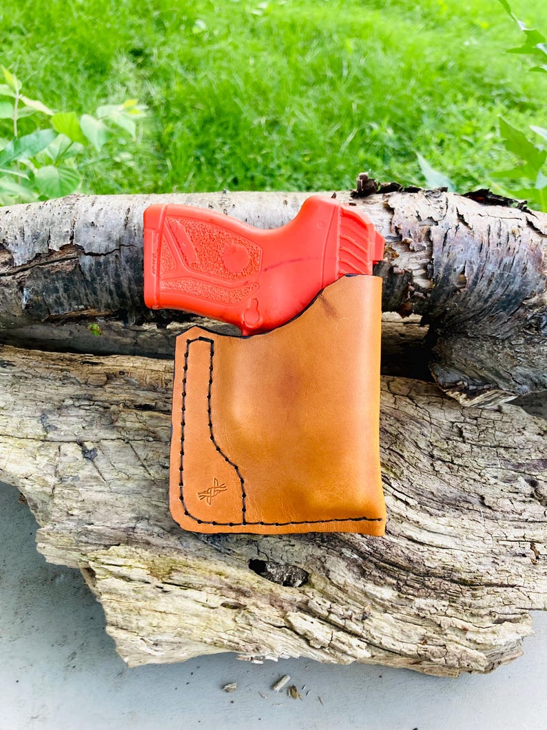 May include: A tan leather holster with black stitching and a red training pistol. The holster has a minimalist design with a stitched logo on the front. The pistol is visible inside the holster.