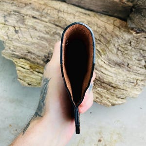 May include: A black leather wallet with a brown interior. The wallet is held open, revealing the inside. The edges are neatly stitched, and the leather has a slightly textured appearance. The wallet is designed to hold cards and cash.