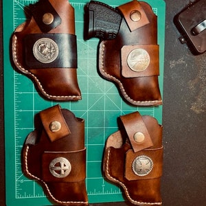 Op de afbeelding: Vier bruine leren holster voor vuurwapens. Twee holster hebben een zilveren cirkelvormig ontwerp met een kruis in het midden. Een holster heeft een zilveren cirkelvormig ontwerp met een vogel in het midden. Een holster heeft een zilveren cirkelvormig ontwerp met een ster in het midden.