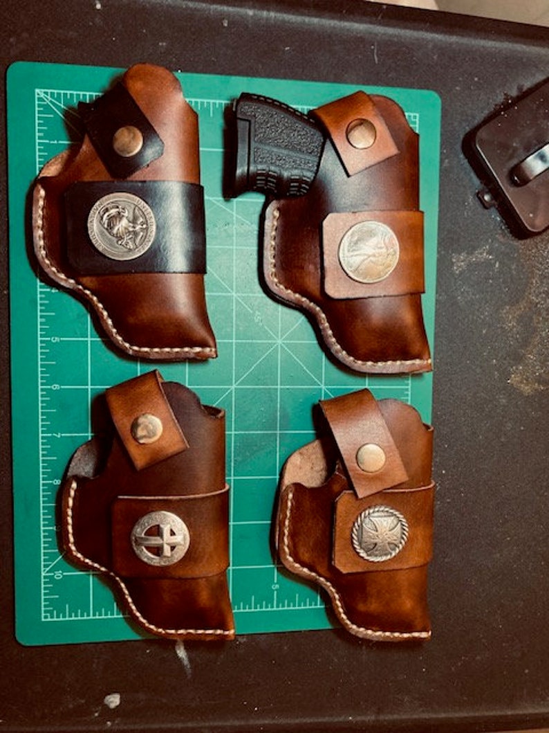 Op de afbeelding: Vier bruine leren holster voor vuurwapens. Twee holster hebben een zilveren cirkelvormig ontwerp met een kruis in het midden. Een holster heeft een zilveren cirkelvormig ontwerp met een vogel in het midden. Een holster heeft een zilveren cirkelvormig ontwerp met een ster in het midden.