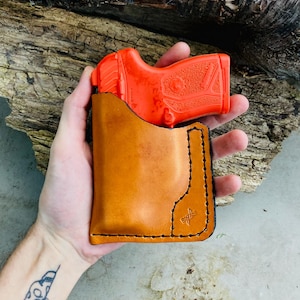 May include: A bright orange training pistol is partially inserted into a tan leather holster. The holster has black stitching and a small logo. The background is a piece of weathered wood.