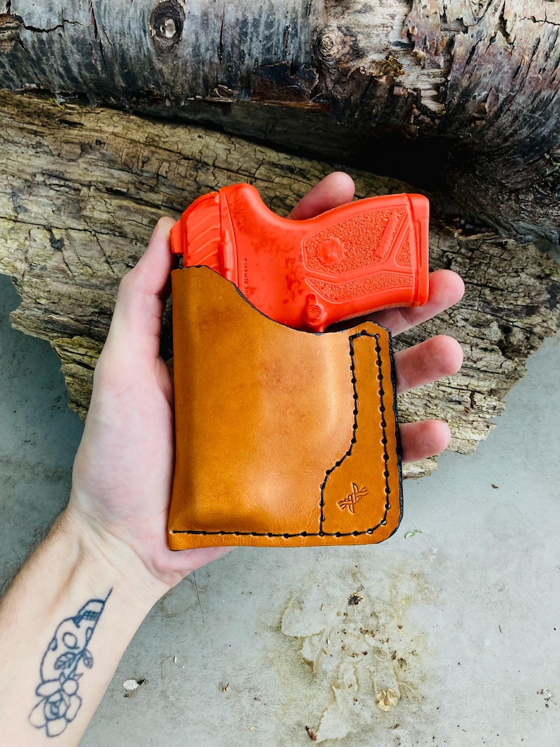 May include: A bright orange training pistol is partially inserted into a tan leather holster. The holster has black stitching and a small logo. The background is a piece of weathered wood.