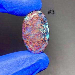 May include: An oval-shaped gemstone with a unique pattern of colors. The stone features a mix of purple, red, and blue hues with a textured appearance. The text "#3" is visible on the right side of the stone. The gemstone is held by a person wearing blue gloves.