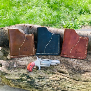 Puede incluir: Tres carteras de cuero en marrón, negro y rojo con un logotipo de oso y una pequeña pistola de plata con mango de madera.