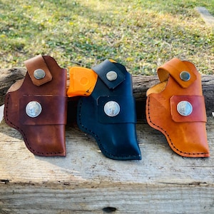 Op de afbeelding: Drie leren holsters in bruin, zwart en oranje, elk met een zilveren knoop en een decoratieve zilveren munt. De holsters worden getoond op een verweerd houten oppervlak, wat hun vakmanschap laat zien.
