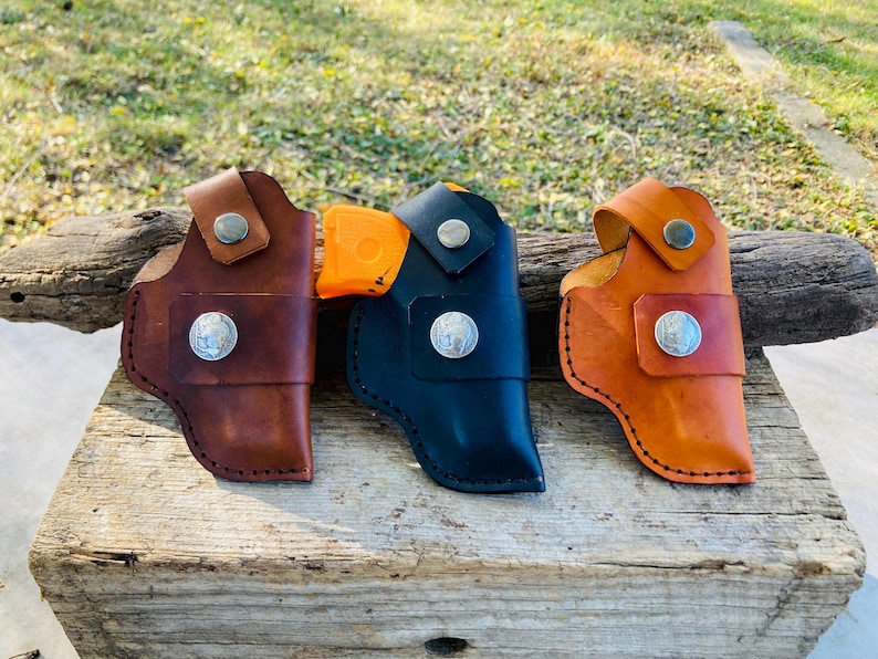 Op de afbeelding: Drie leren holsters in bruin, zwart en oranje, elk met een zilveren knoop en een decoratieve zilveren munt. De holsters worden getoond op een verweerd houten oppervlak, wat hun vakmanschap laat zien.