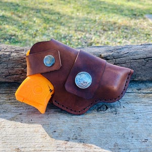 Op de afbeelding: Een bruin leren holster met een zilveren knoop en een zilveren munt met een bizon. Een oranje pistool is gedeeltelijk zichtbaar. De holster ligt op een verweerd houten oppervlak.