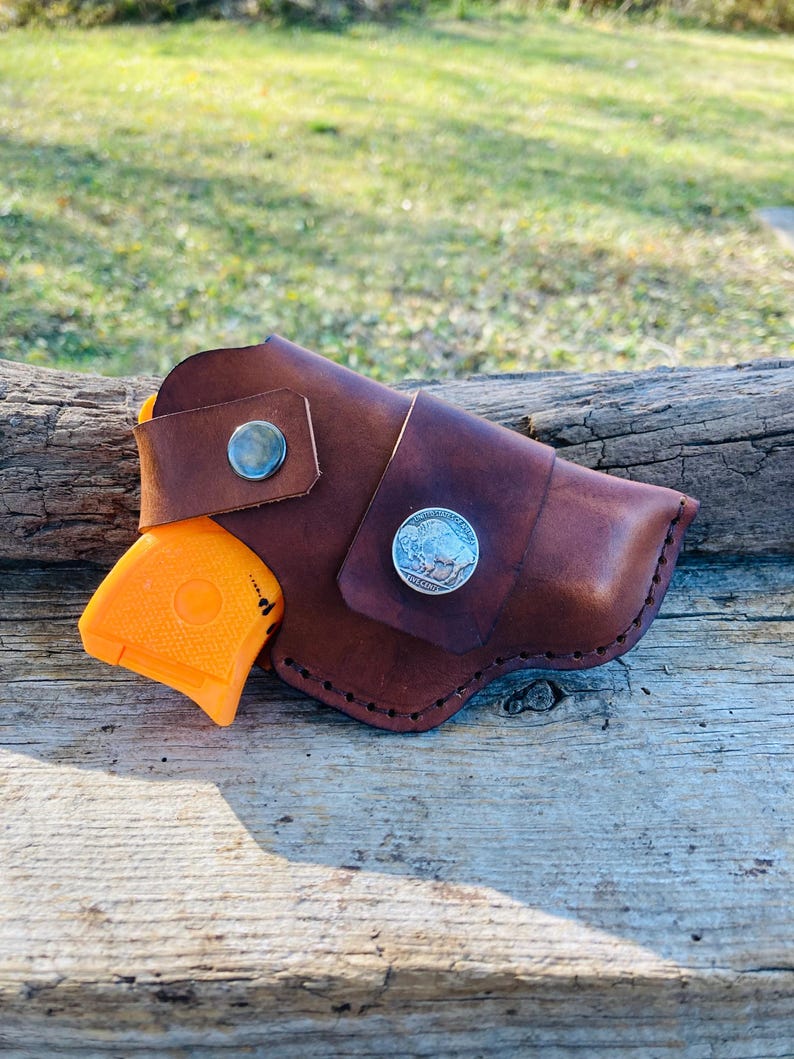 Op de afbeelding: Een bruin leren holster met een zilveren knoop en een zilveren munt met een bizon. Een oranje pistool is gedeeltelijk zichtbaar. De holster ligt op een verweerd houten oppervlak.
