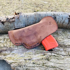 K&ouml;nnte beinhalten: Ein braunes Lederholster mit einem gen&auml;hten Rand und einem kleinen Logo auf der R&uuml;ckseite. Das Holster h&auml;lt eine leuchtend orangefarbene Pistole.