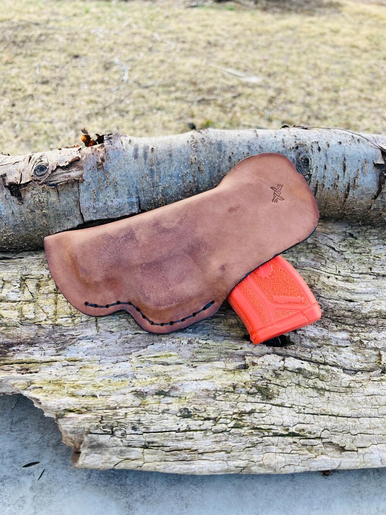 K&ouml;nnte beinhalten: Ein braunes Lederholster mit einem gen&auml;hten Rand und einem kleinen Logo auf der R&uuml;ckseite. Das Holster h&auml;lt eine leuchtend orangefarbene Pistole.