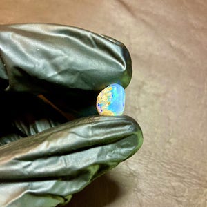Puede incluir: Una vibrante piedra preciosa de ópalo, que muestra una mezcla de colores azul, dorado y blanco. La piedra se sostiene entre dedos enguantados, mostrando sus patrones y texturas únicos. El ópalo es de forma pequeña e irregular, con una superficie pulida.