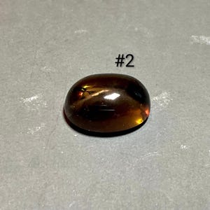 May include: A polished, oval-shaped gemstone with a rich, amber color. The gem has a smooth, rounded surface and reflects light, creating highlights. The number "#2" is printed in white above the gem.