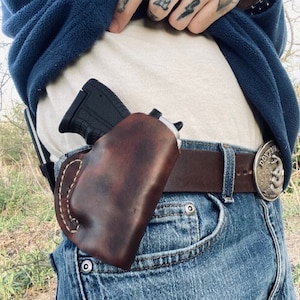 Könnte beinhalten: Ein braunes Lederholster mit einer schwarzen Pistole darin. Das Holster ist an einem braunen Ledergürtel mit einer silbernen Schnalle befestigt, auf der "Don't Tread On Me" steht.