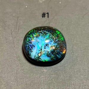 Puede incluir: Ópalo ovalado pulido con colores vibrantes. La gema presenta una base de tonos azules y verdes, con destellos dorados e inclusiones negras. El texto "#1" es visible encima del ópalo.