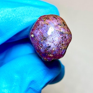 Pode incluir: Uma pedra preciosa bruta e não polida com um tom magenta e roxo profundo, apresentando inclusões naturais na superfície, veios minerais escuros e uma estrutura cristalina semitranslúcida de forma arredondada e irregular com um reflexo brilhante.