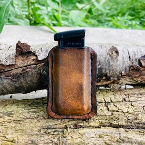 Może przedstawiać: Brązowe skórzane etui na magazynek z włożonym czarnym magazynkiem. Etui wykonane jest ze skóry postarzanej i ma szyte krawędzie. Tekst "MADE IN USA" jest widoczny na magazynku.