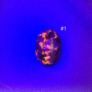 May include: An oval-shaped stone cabochon displaying a mottled pattern of vibrant orange and deep purple under ultraviolet light, with the text #1 positioned to the right of the stone. The surface features dark speckles and reflective highlights.