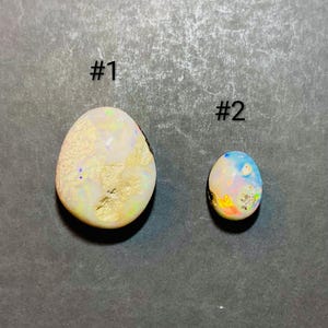 Puede incluir: Dos ópalos sobre una superficie gris oscuro. El ópalo más grande, etiquetado como #1, es de forma ovalada con una base crema y destellos iridiscentes de color. El ópalo más pequeño, etiquetado como #2, también es ovalado, mostrando tonos azules, rosas y amarillos.