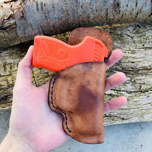K&ouml;nnte beinhalten: Ein braunes Lederholster f&uuml;r eine kleine, orangefarbene Pistole. Das Holster hat einen gen&auml;hten Rand und wird in der Hand einer Person gehalten.