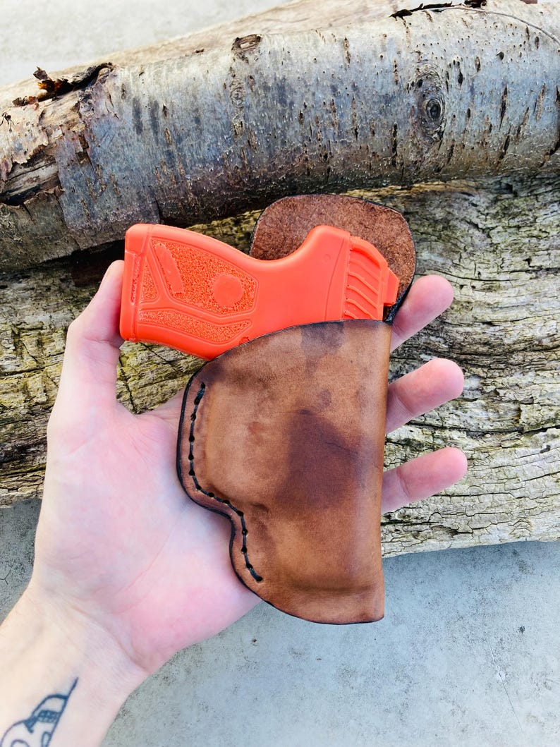 K&ouml;nnte beinhalten: Ein braunes Lederholster f&uuml;r eine kleine, orangefarbene Pistole. Das Holster hat einen gen&auml;hten Rand und wird in der Hand einer Person gehalten.