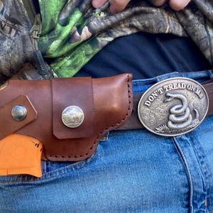 Op de afbeelding: Een bruin leren holster met een zilveren knoop en een zilveren munt is bevestigd aan de spijkerbroek van een persoon. Een oranje object is gedeeltelijk zichtbaar in de holster. Een zilveren gesp met de woorden "DON'T TREAD ON ME" en een opgerolde slang is ook zichtbaar.