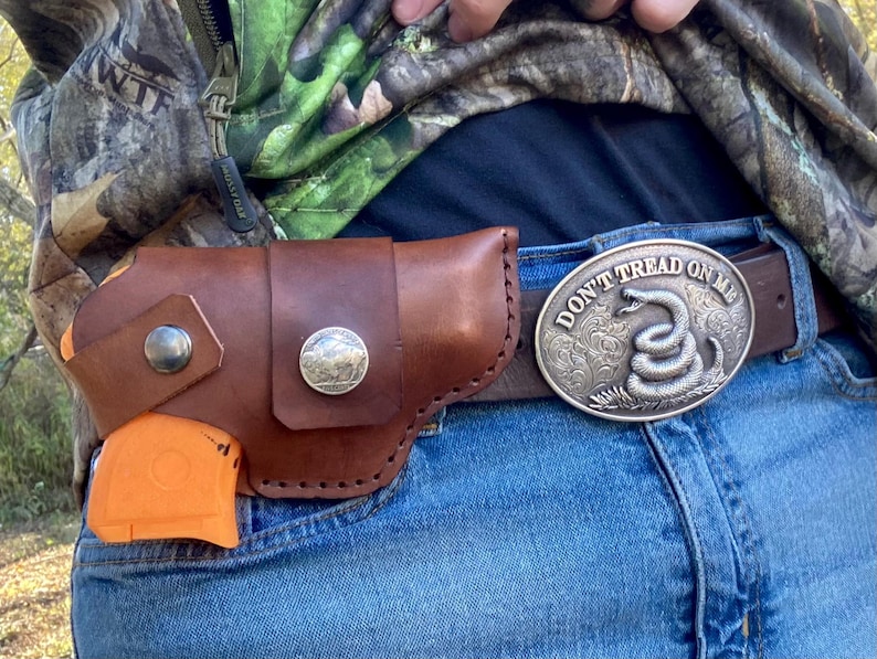 Op de afbeelding: Een bruin leren holster met een zilveren knoop en een zilveren munt is bevestigd aan de spijkerbroek van een persoon. Een oranje object is gedeeltelijk zichtbaar in de holster. Een zilveren gesp met de woorden "DON'T TREAD ON ME" en een opgerolde slang is ook zichtbaar.