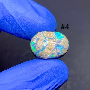 Puede incluir: Una piedra preciosa de ópalo ovalada con una mezcla de colores, incluyendo azul, verde y dorado, es sostenida por una mano enguantada. El ópalo tiene un patrón y textura únicos. El número "#4" es visible en la imagen.