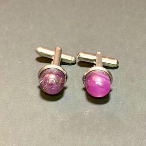 May include: A pair of cufflinks with silver-colored metal hardware. Each cufflink features a round, polished, pinkish-purple stone. The cufflinks are displayed on a gray surface.