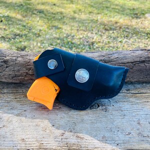 Op de afbeelding: Zwarte leren holster met een zilveren knoop en een zilveren munt detail. Een oranje pistool is gedeeltelijk zichtbaar. De holster is ontworpen om een klein vuurwapen te bevatten. De achtergrond is een houten oppervlak.