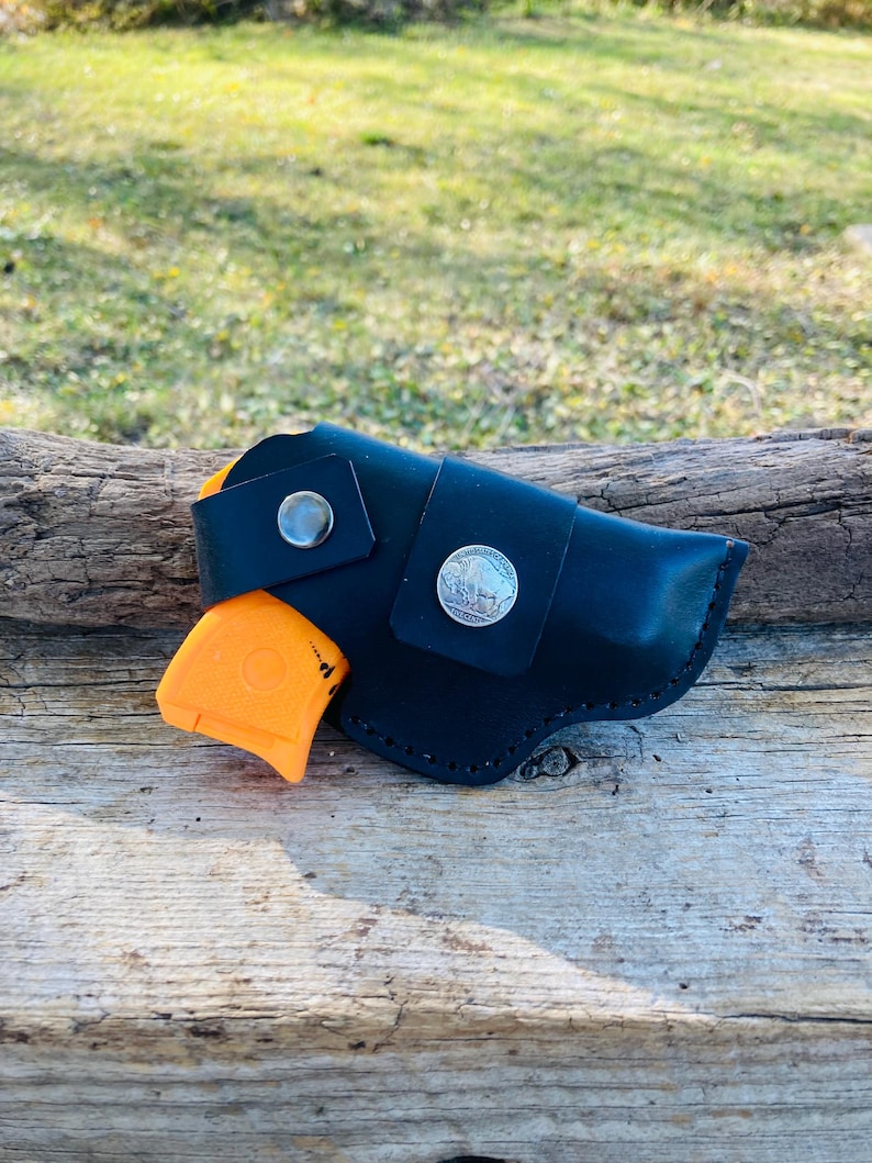 Op de afbeelding: Zwarte leren holster met een zilveren knoop en een zilveren munt detail. Een oranje pistool is gedeeltelijk zichtbaar. De holster is ontworpen om een klein vuurwapen te bevatten. De achtergrond is een houten oppervlak.