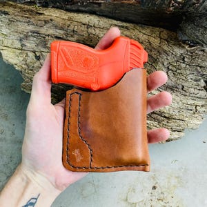 May include: A bright orange toy gun is partially inserted into a brown leather holster. The holster is hand-stitched with black thread and features a decorative emblem. The background includes a weathered wooden log.