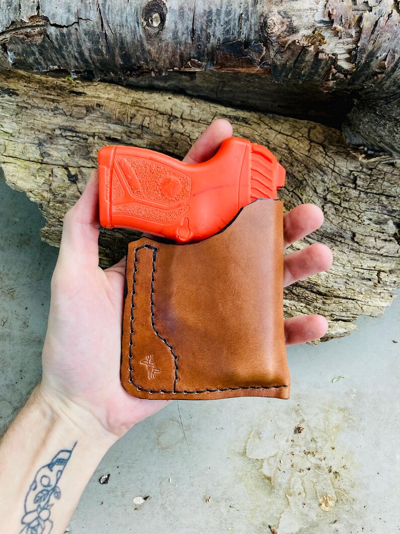 May include: A bright orange toy gun is partially inserted into a brown leather holster. The holster is hand-stitched with black thread and features a decorative emblem. The background includes a weathered wooden log.