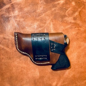 Op de afbeelding: Een bruine leren holster met een zwarte leren riem en een zwart pistool erin.