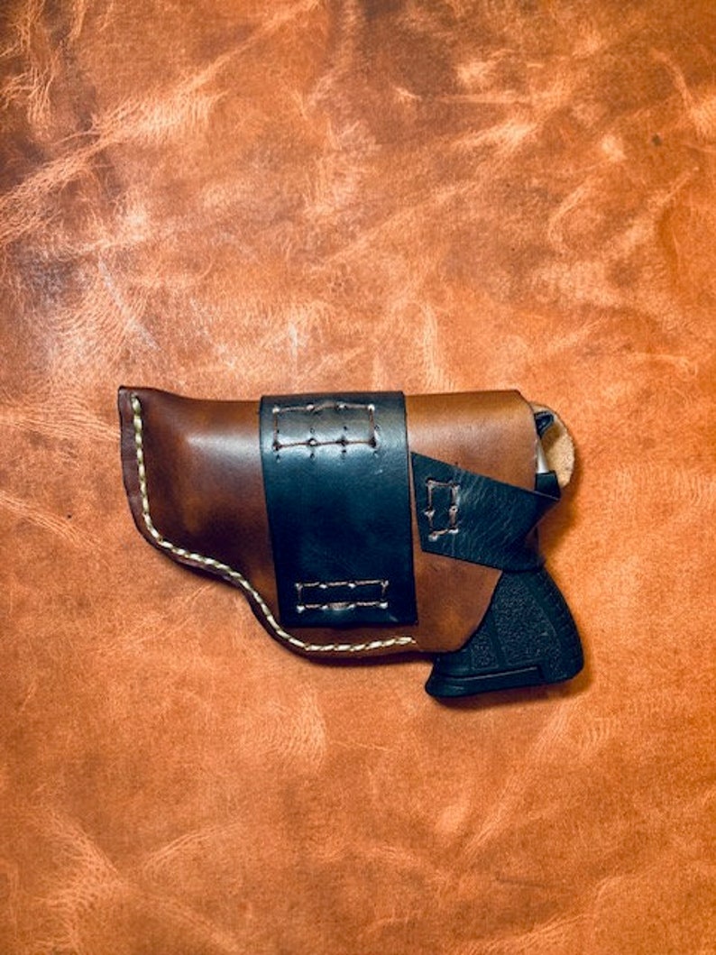 Op de afbeelding: Een bruine leren holster met een zwarte leren riem en een zwart pistool erin.