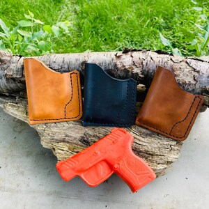 May include: Four leather wallets in tan, black, and brown, with black stitching and a small cross detail. An orange toy gun is placed below the wallets. The items are displayed on a piece of wood against green grass.