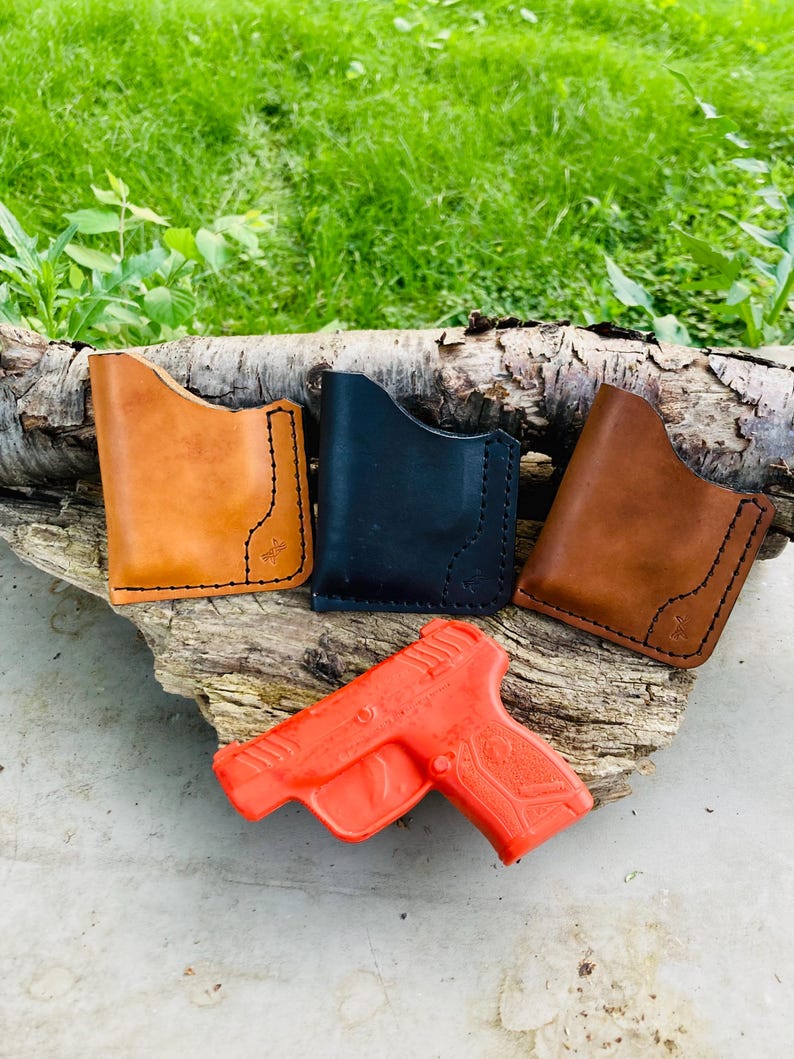 May include: Four leather wallets in tan, black, and brown, with black stitching and a small cross detail. An orange toy gun is placed below the wallets. The items are displayed on a piece of wood against green grass.