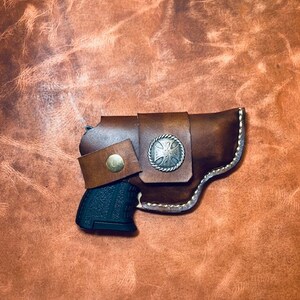 Op de afbeelding: Een bruine leren holster met een zilveren concho en een zwart pistool erin.